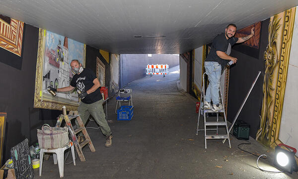 Bild vergrößern: Künstler links sprüht das Motiv Kreuztor in einem Rahmen an die Wand, Künstler rechts auf einer Leiter mit Spraydose in der Hand